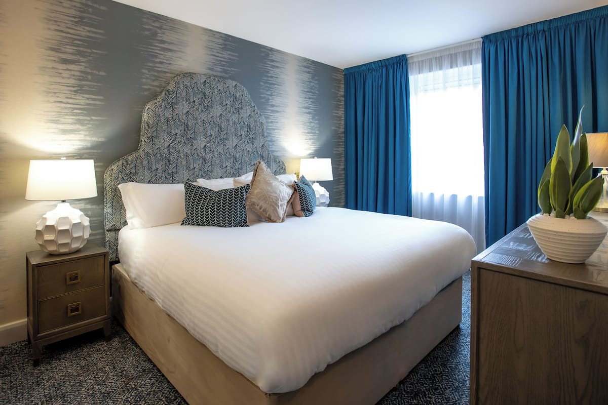 Een netjes opgemaakt bed met een bed hoofd bord met patroon, twee nachtkastjes met lampen, een groot raam met blauwe gordijnen, en een potplant op een bijzettafeltje.