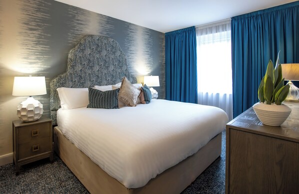 A neatly made bed with a patterned headboard, two bedside tables with lamps, a large window with blue curtains, and a potted plant on a side table.