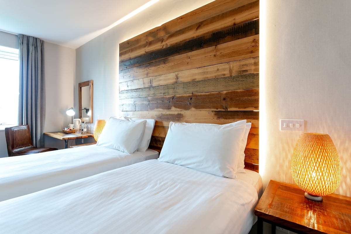 Hotelkamer met twee bedden, een houten hoofdeinde, een bureau met lamp en een raam met gordijnen.