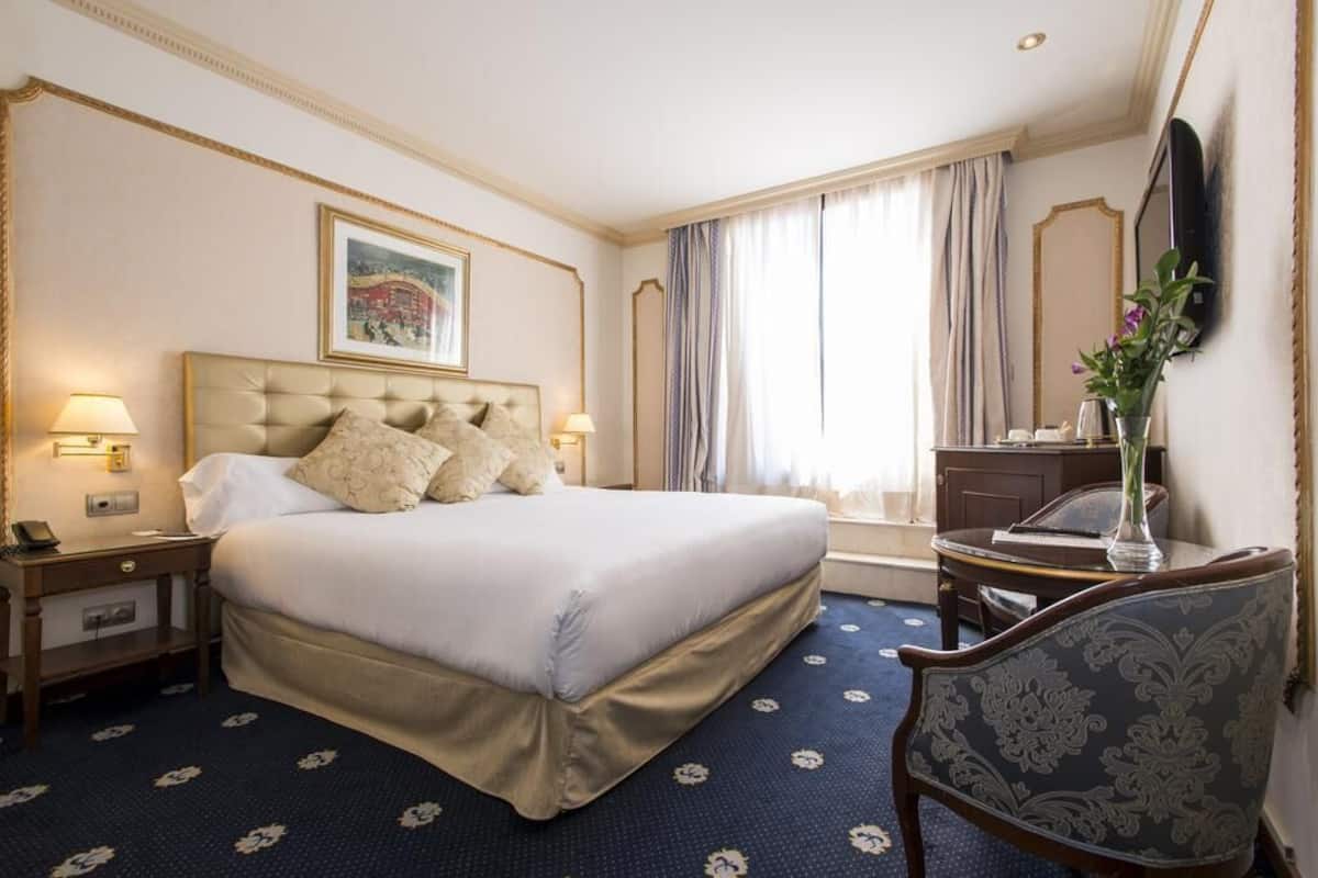 Une chambre d’hôtel avec un grand lit, des lampes de chevet, un bureau, une chaise, un miroir et une fenêtre avec des rideaux.