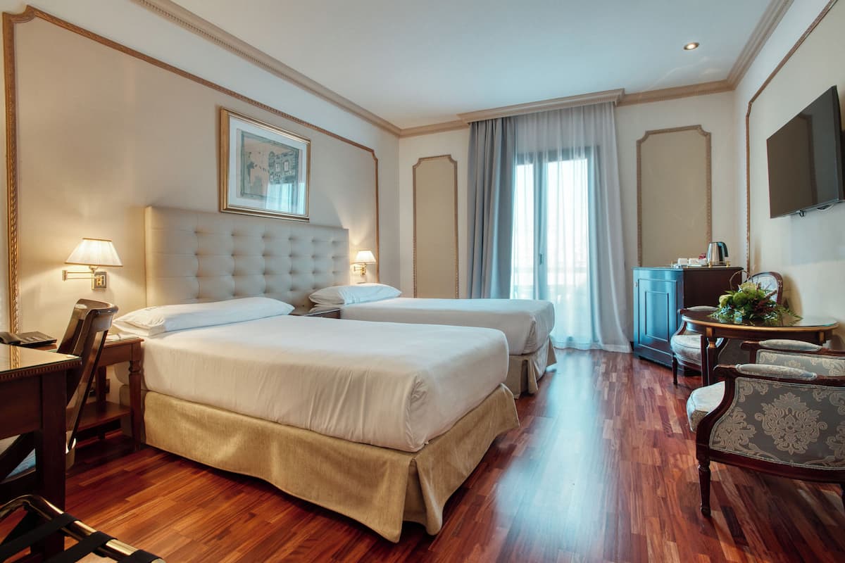 A hotel room with two beds, a desk, a chair, a television, and a window with curtains.