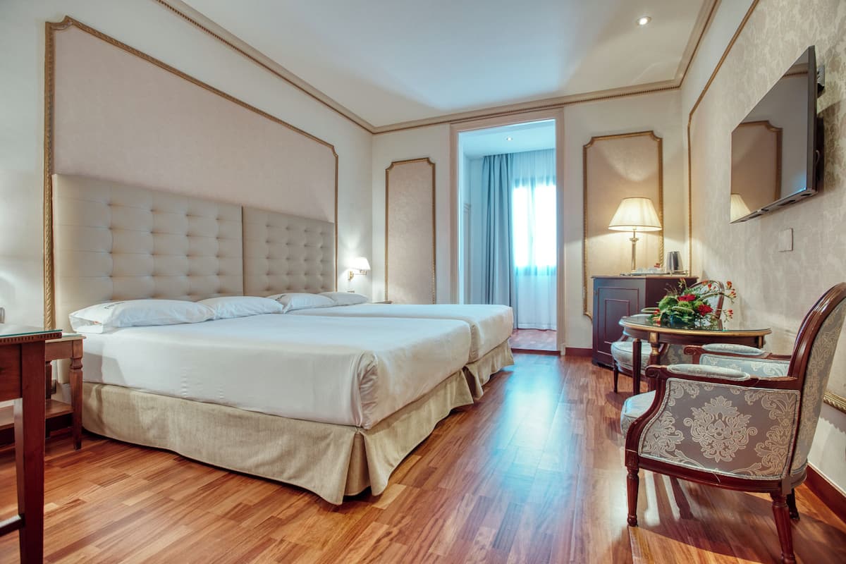 A hotel room with a large bed, a desk, a chair, a TV, and a lamp.