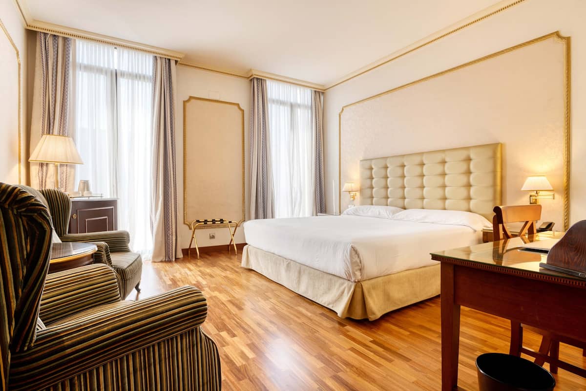 A hotel room with a large bed, a wooden desk, a chair, a sofa, and a lamp.
