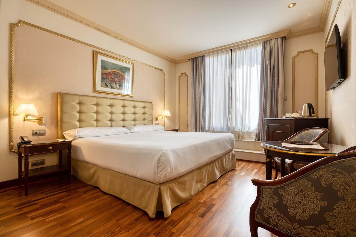A hotel room with a large bed, two bedside tables with lamps, a desk with a chair, a television, and a window with curtains.