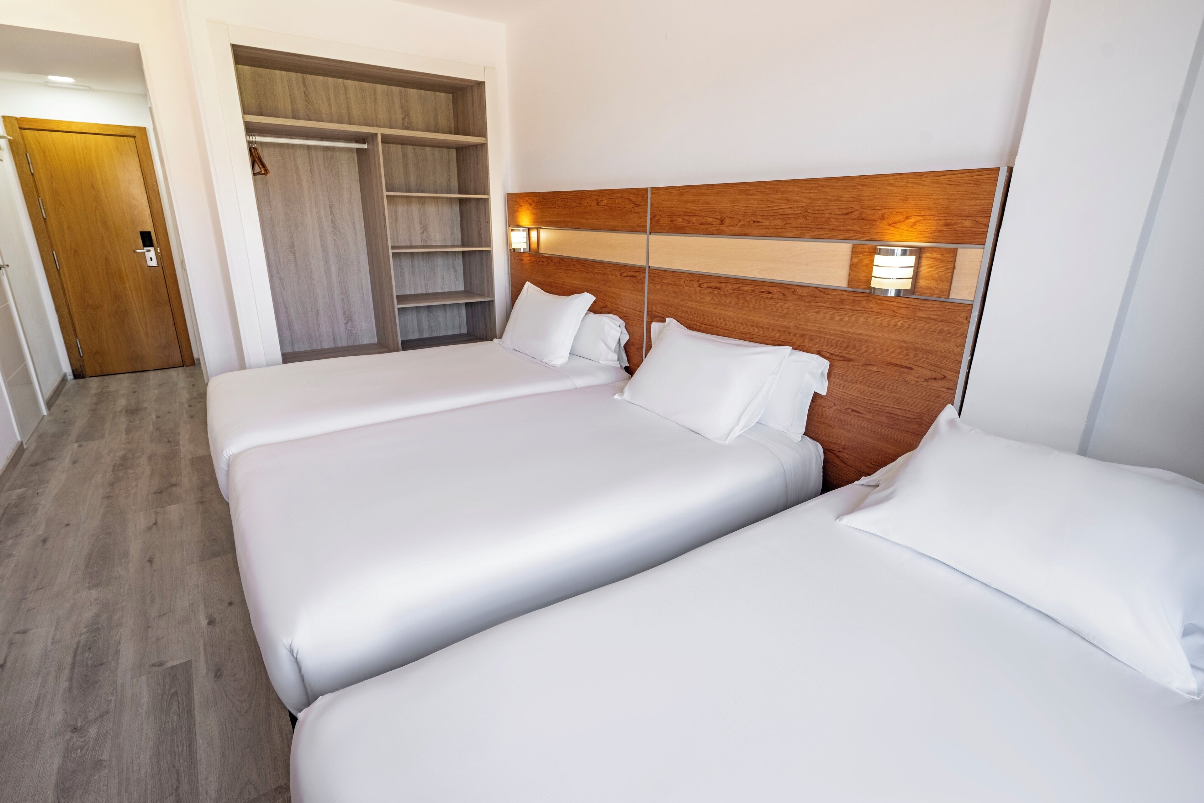 A hotel room with two beds, a wooden headboard, a wardrobe, and a bedside lamp.