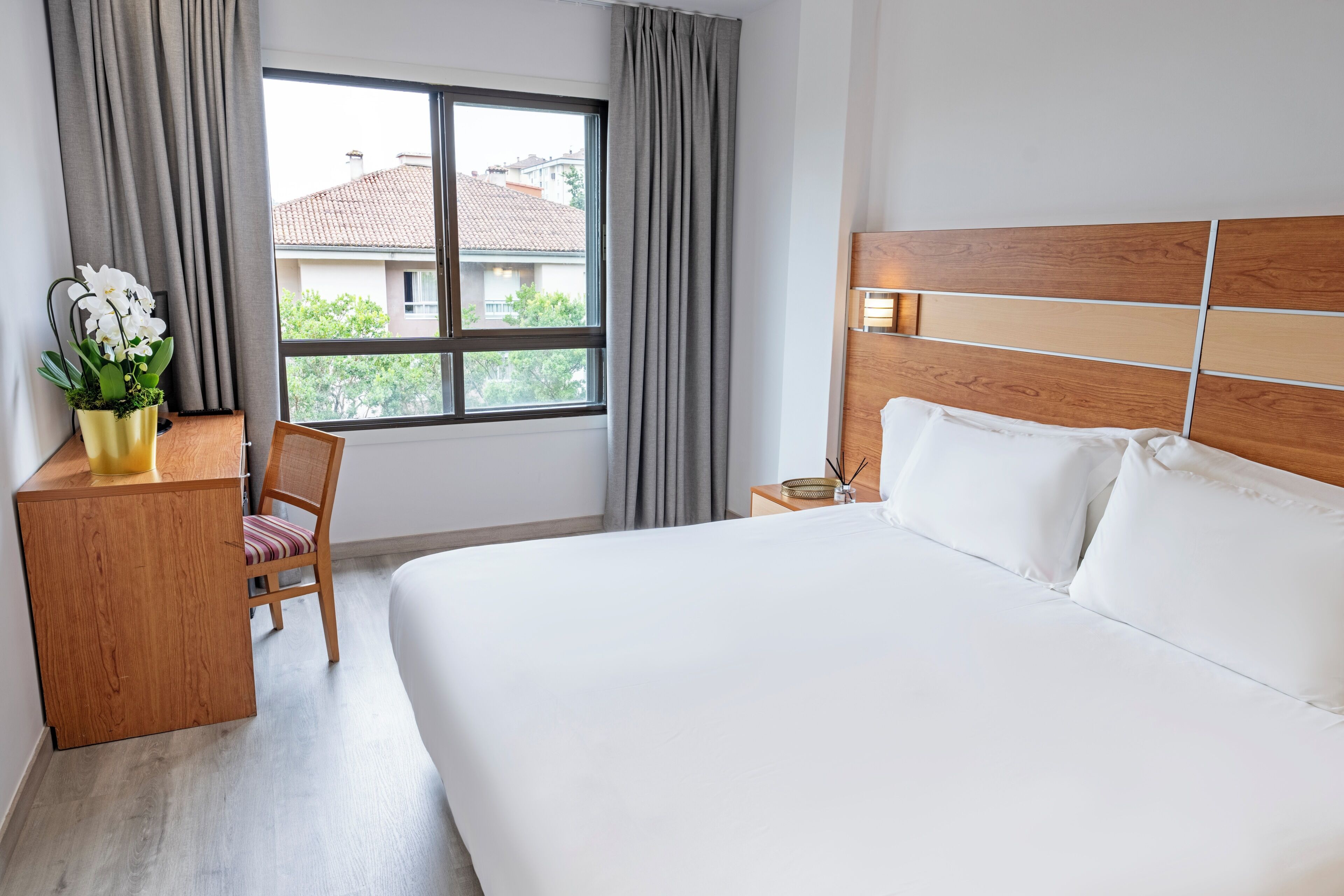 A hotel room with a large bed, a wooden headboard, a desk with a chair, a window with curtains, and a view of buildings outside.