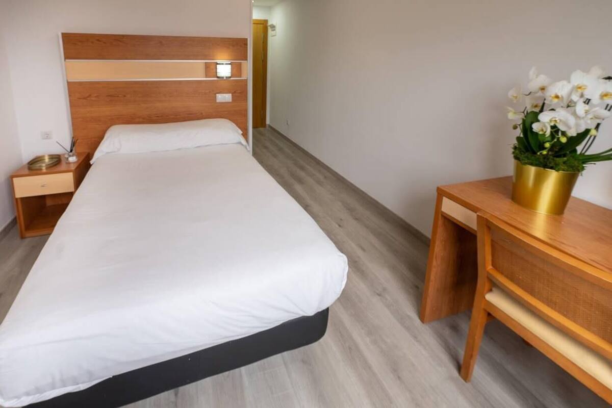 Ein Hotelzimmer mit einem hölzernen Bett, einem Nachttisch mit Lampe, einem kleinen Beistelltisch und einem Schreibtisch mit Blumen in einer Vase.
