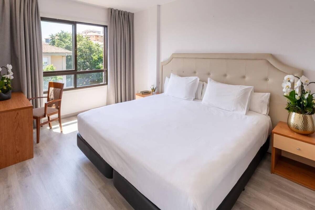 Ein Hotelzimmer mit einem großen Bett, einem Holztisch, einem Stuhl und einem Vorhangfenster.