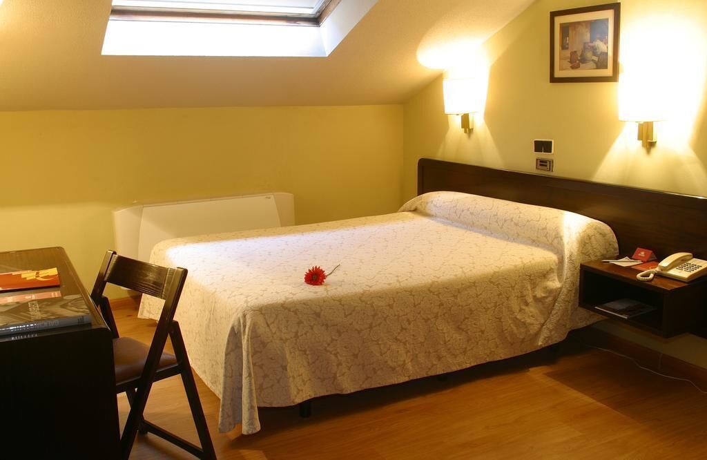 A hotel room with a bed, a desk with a chair, a bedside table with a phone, and a picture on the wall.