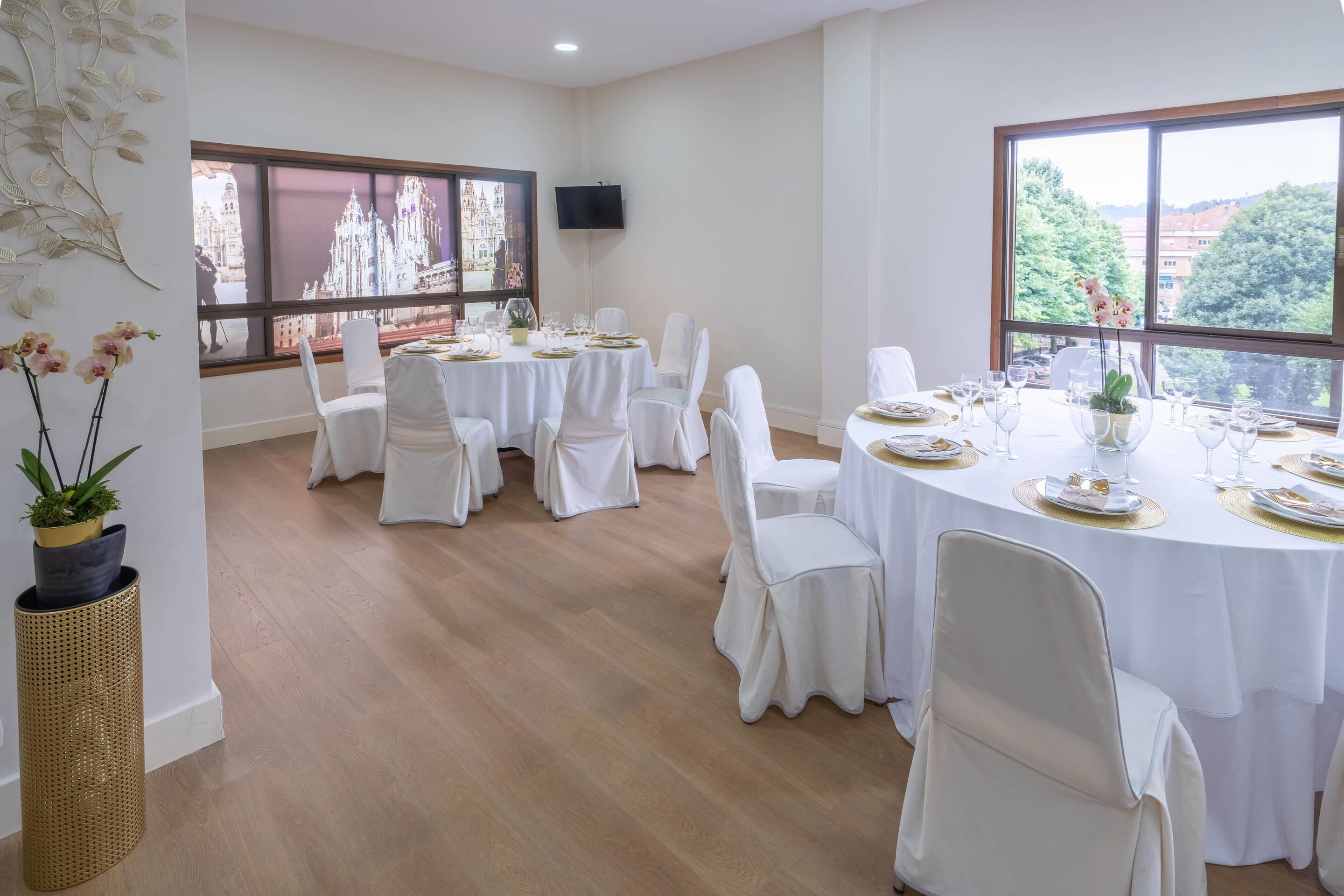 Uma área de refeições com mesas redondas preparadas para uma refeição, cadeiras brancas e janelas amplas com vista para edifícios e vegetação.