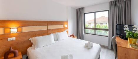 A hotel room with a large bed, a wooden headboard, a desk with a computer, and a window with curtains.