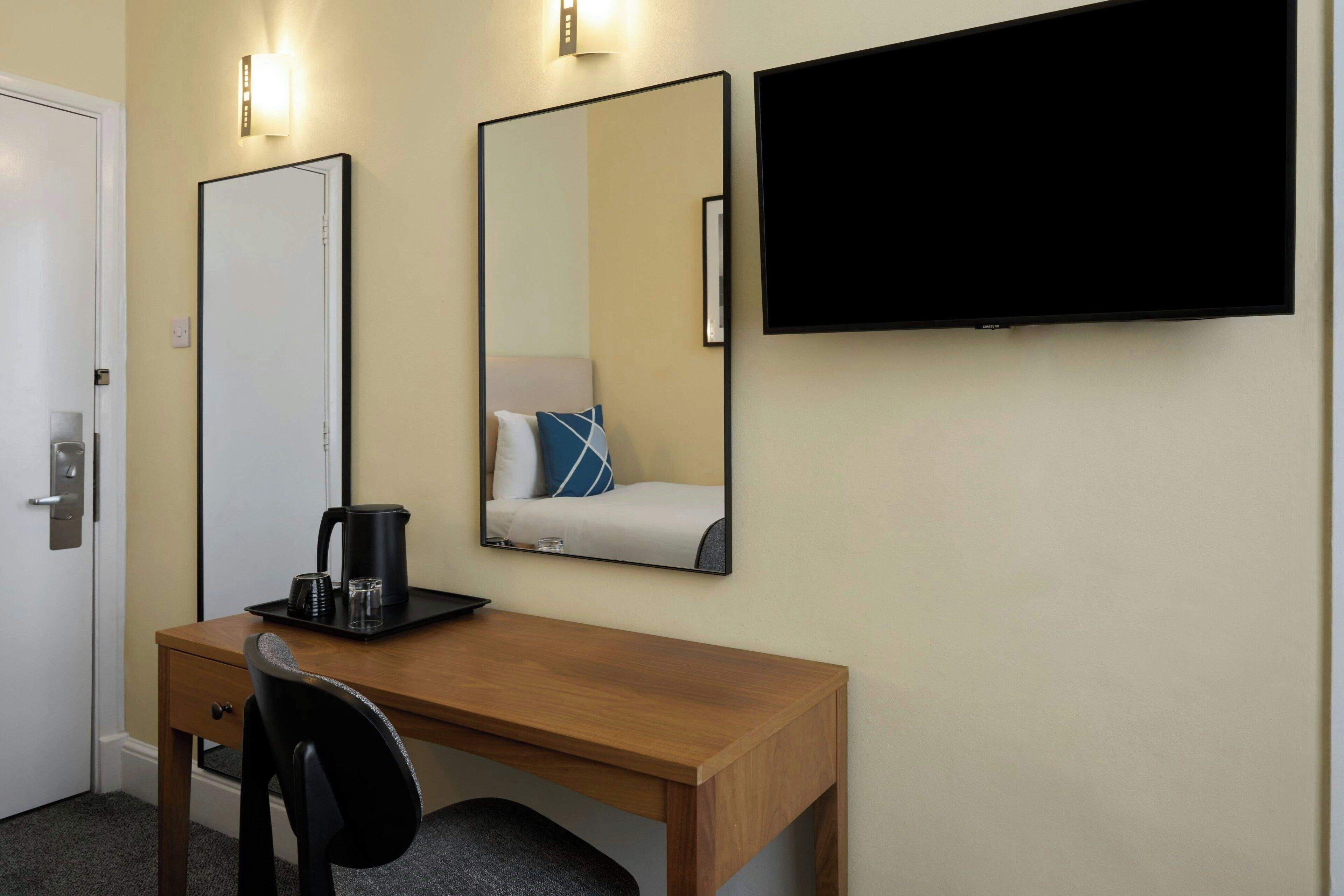 A hotel room with a wooden desk, a chair, a coffee maker, two mirrors, and a flat-screen TV.