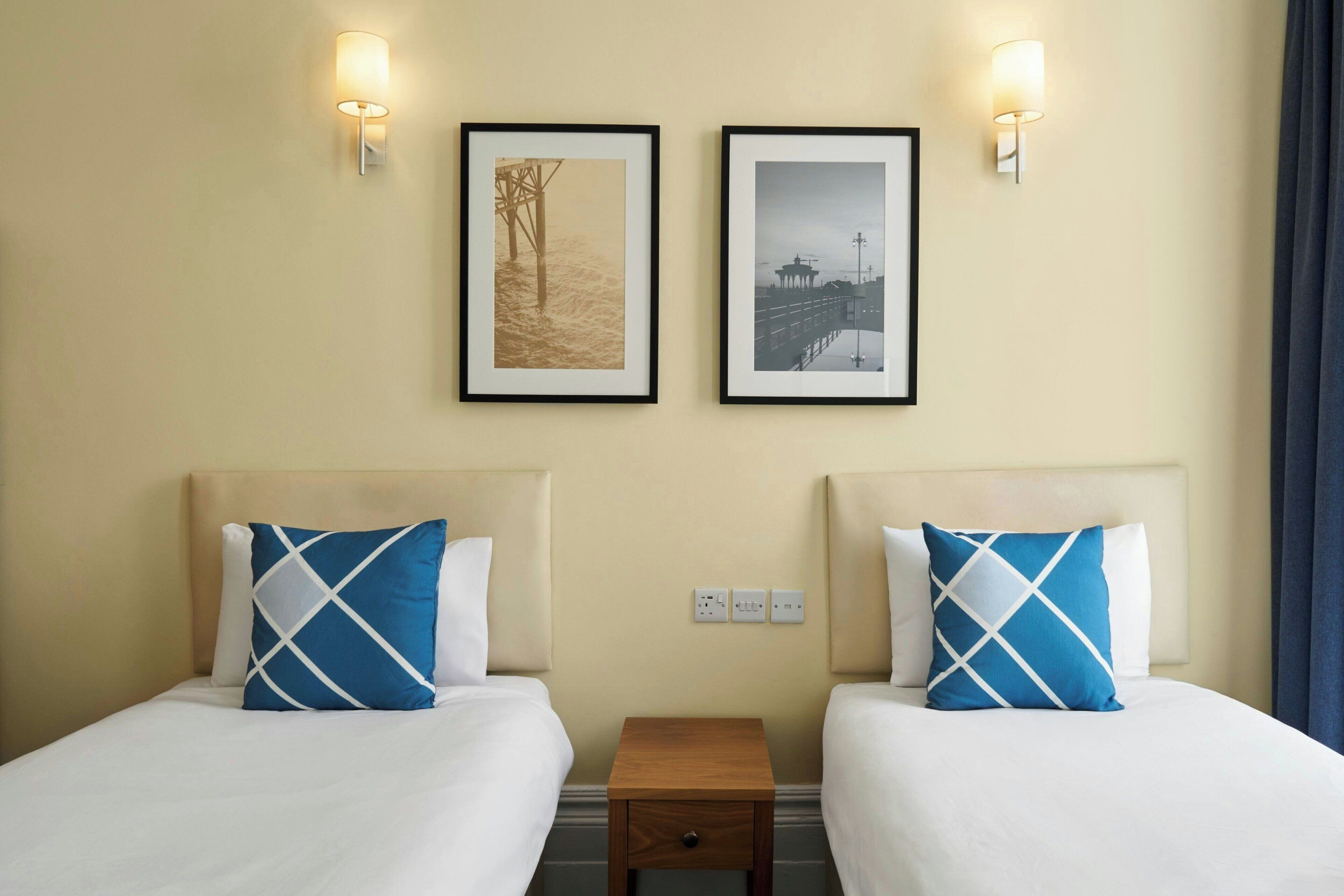 A hotel room with two beds, each with a pillow and a nightstand. There are two framed pictures on the wall, and the room has a neutral color scheme.