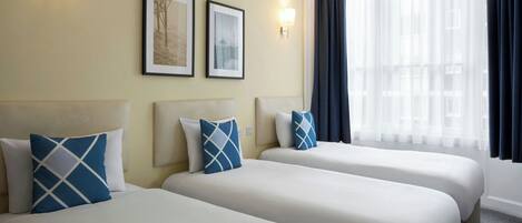Habitación de hotel con dos camas, cada una con cubrecama blanco y almohadas azules, un ventanal con cortinas traslúcidas y lámparas fijadas a la pared.