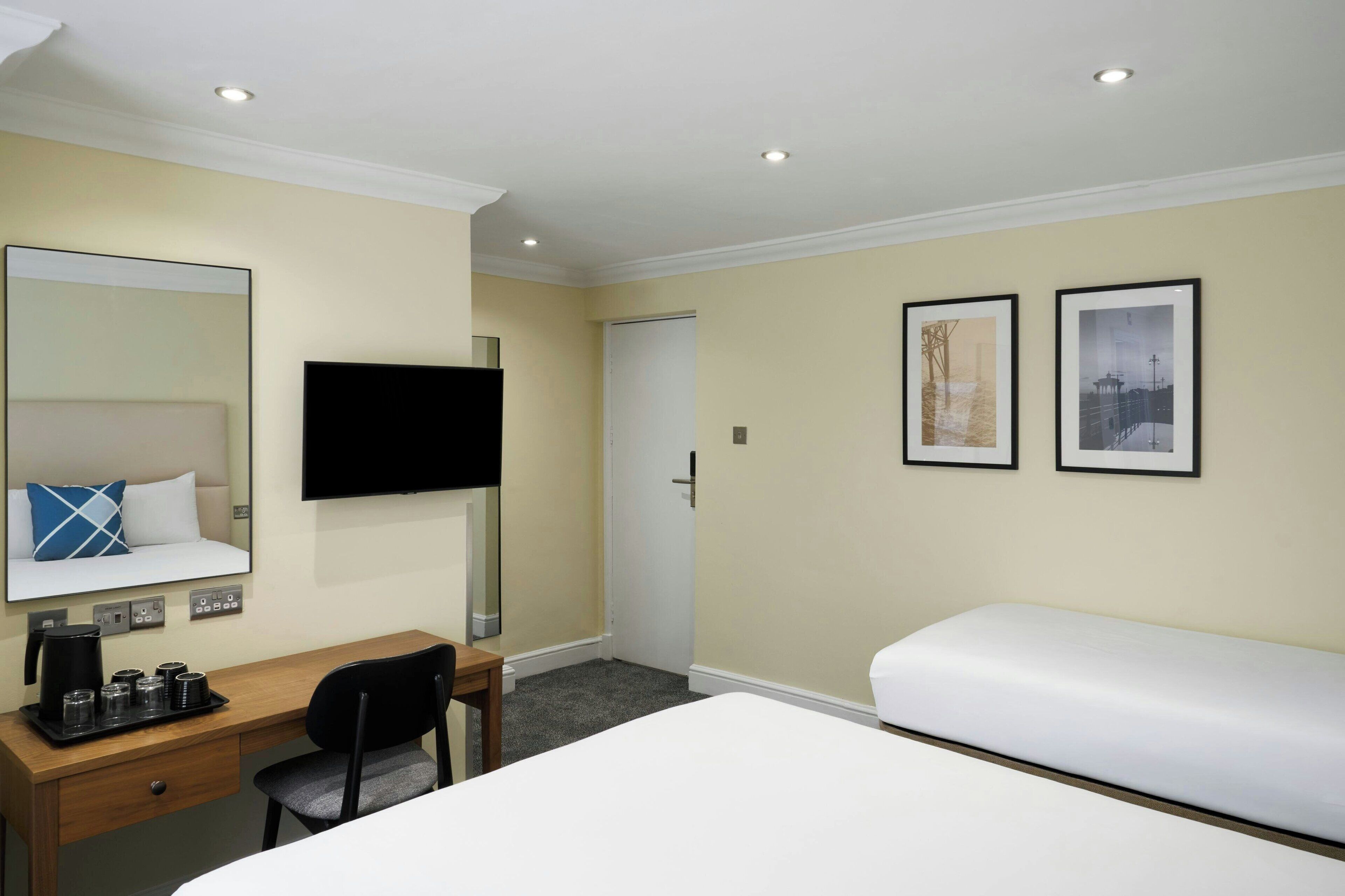 A hotel room with a bed, a desk with a chair, a flat-screen TV, a coffee station, and two framed pictures on the wall.
