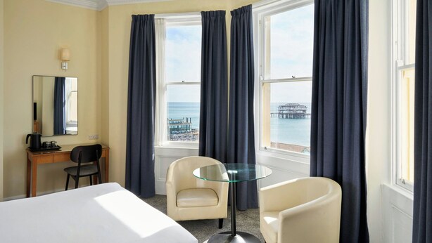 Une chambre d’hôtel comprenant un lit, deux chaises, un bureau, une machine à café, une table en verre et une grande fenêtre avec des rideaux.