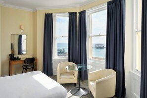 Habitación de hotel con una cama, dos sillas, un escritorio, una cafetera, una mesita de cristal y un ventanal grande con cortinas.