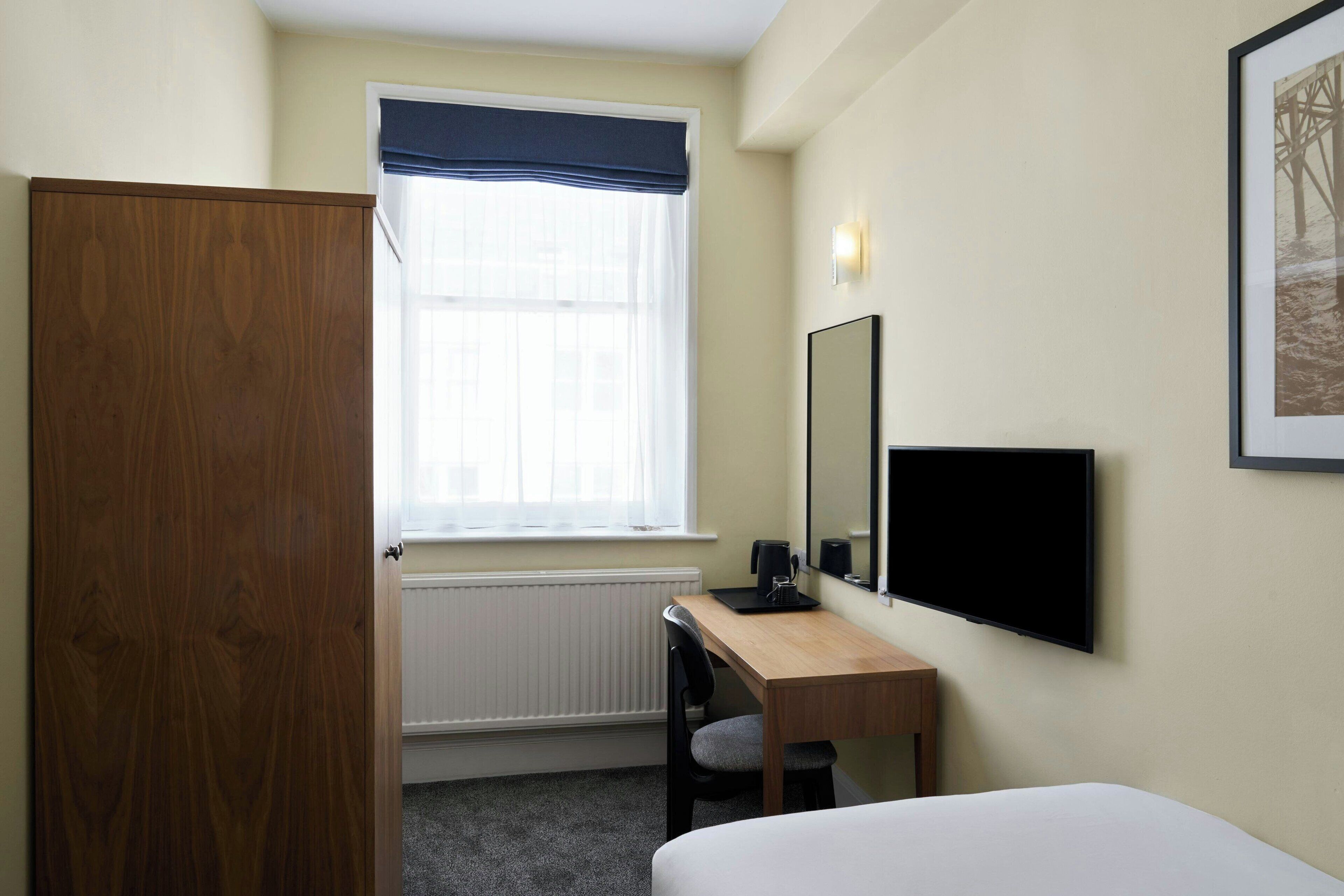 A hotel room with a wooden wardrobe, a desk with a chair, a bed, a window with a blue valance, a wall-mounted TV, and a framed picture.