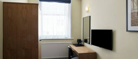A hotel room with a wooden wardrobe, a desk with a chair, a bed, a window with a blue valance, a wall-mounted TV, and a framed picture.