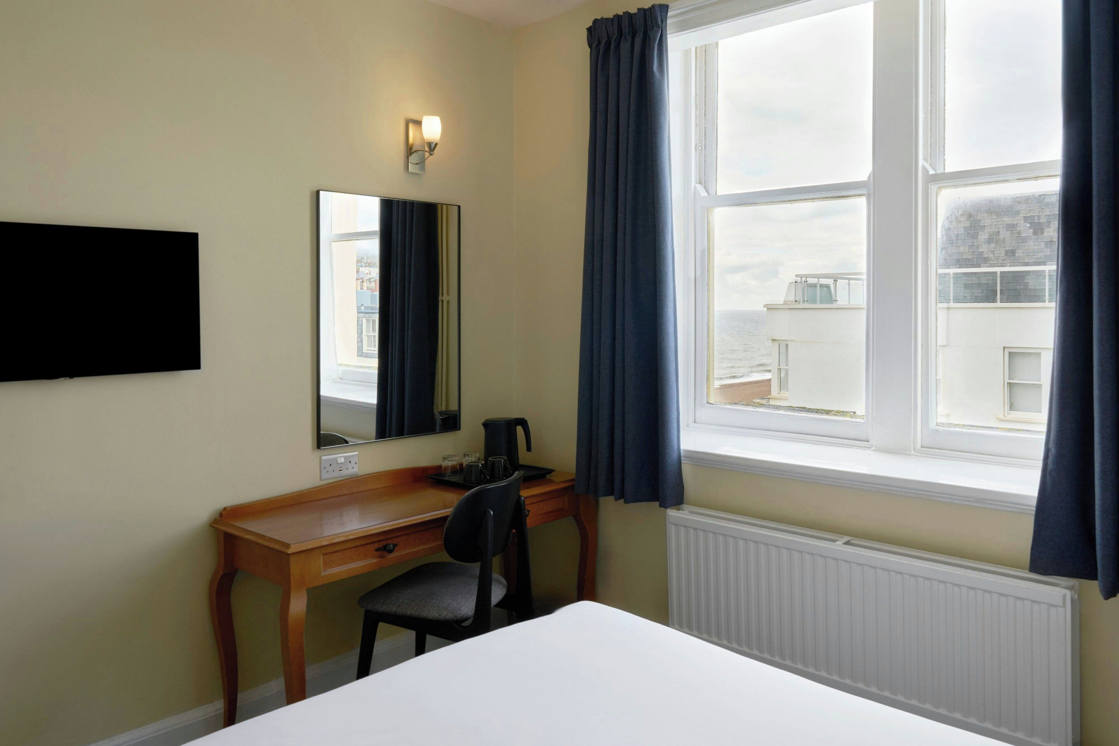 A hotel room with a bed, a desk with a chair, a flat-screen TV, a window with curtains, and a view of buildings outside.