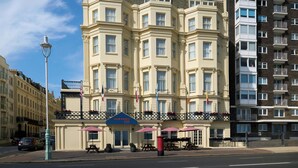Un hôtel à plusieurs étages, de style architectural classique, qui se distingue par son entrée imposante, surmontée d’un auvent bleu, et par son espace extérieur aménagé pour accueillir les clients.