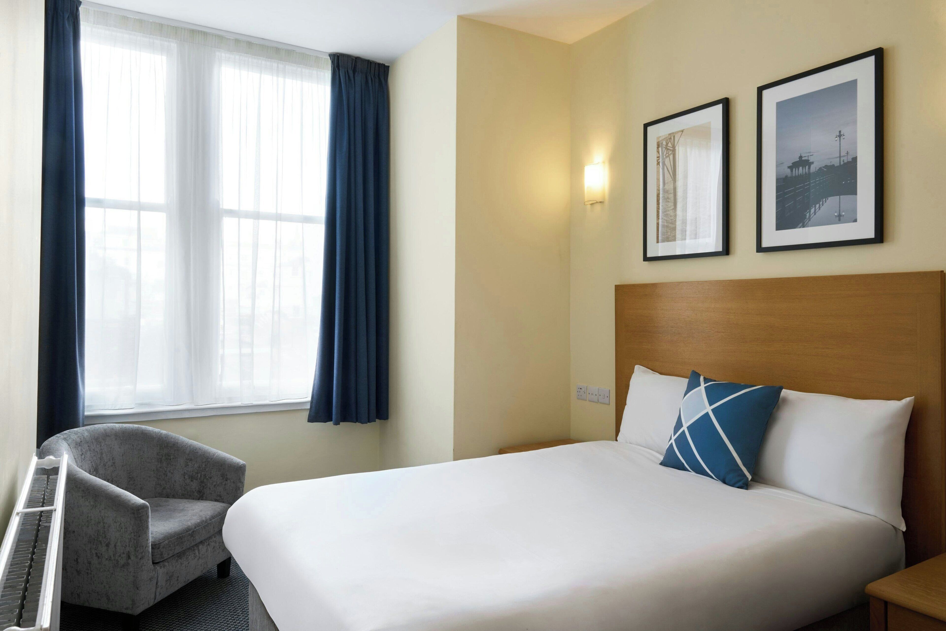 A hotel room with a bed, a chair, a window with curtains, and two framed pictures on the wall.