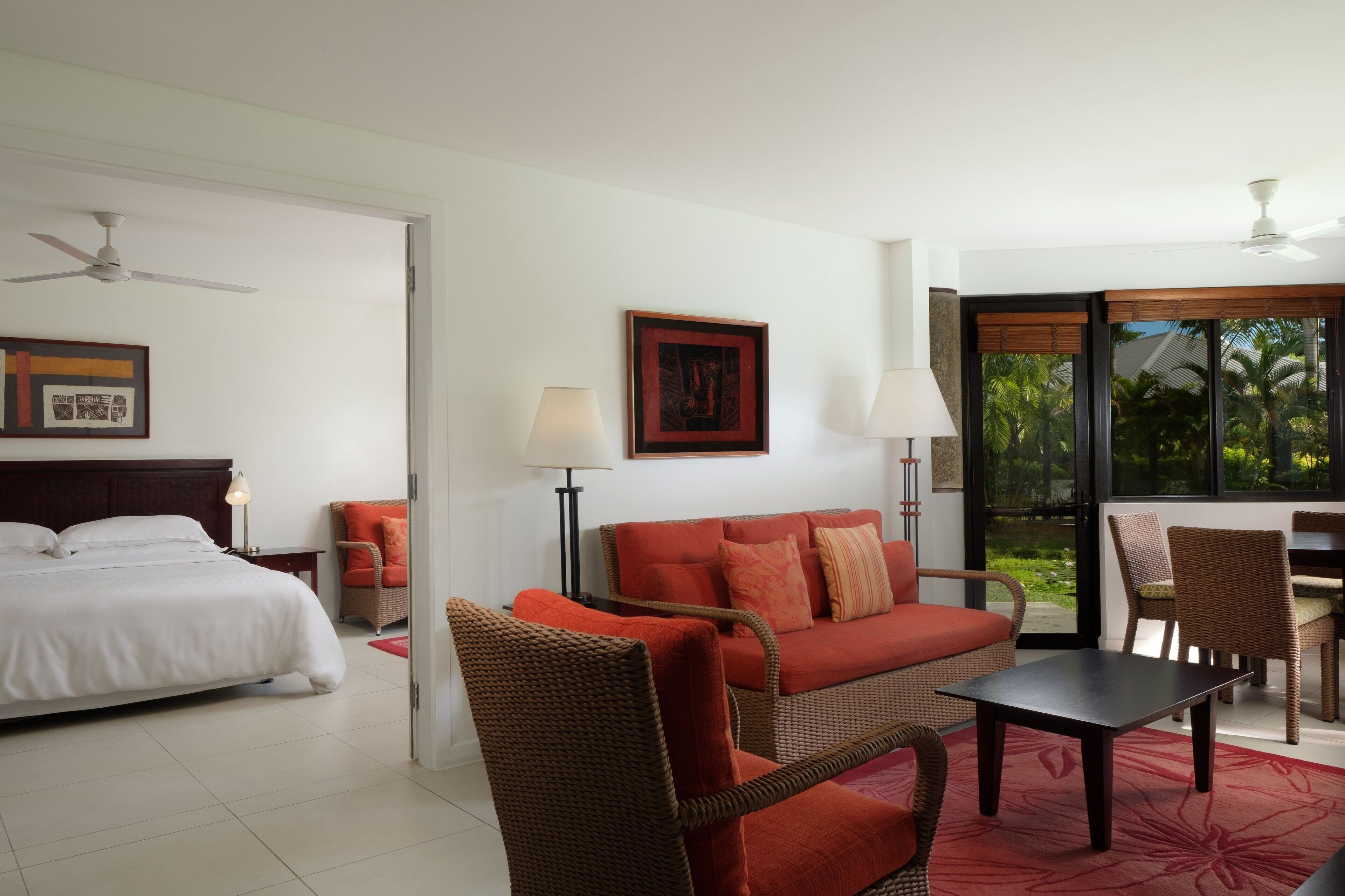 A hotel room with a bed, a red sofa, a dining table, and a chair. There is a window with a view of palm trees.