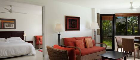 A hotel room with a bed, a red sofa, a dining table, and a chair. There is a window with a view of palm trees.