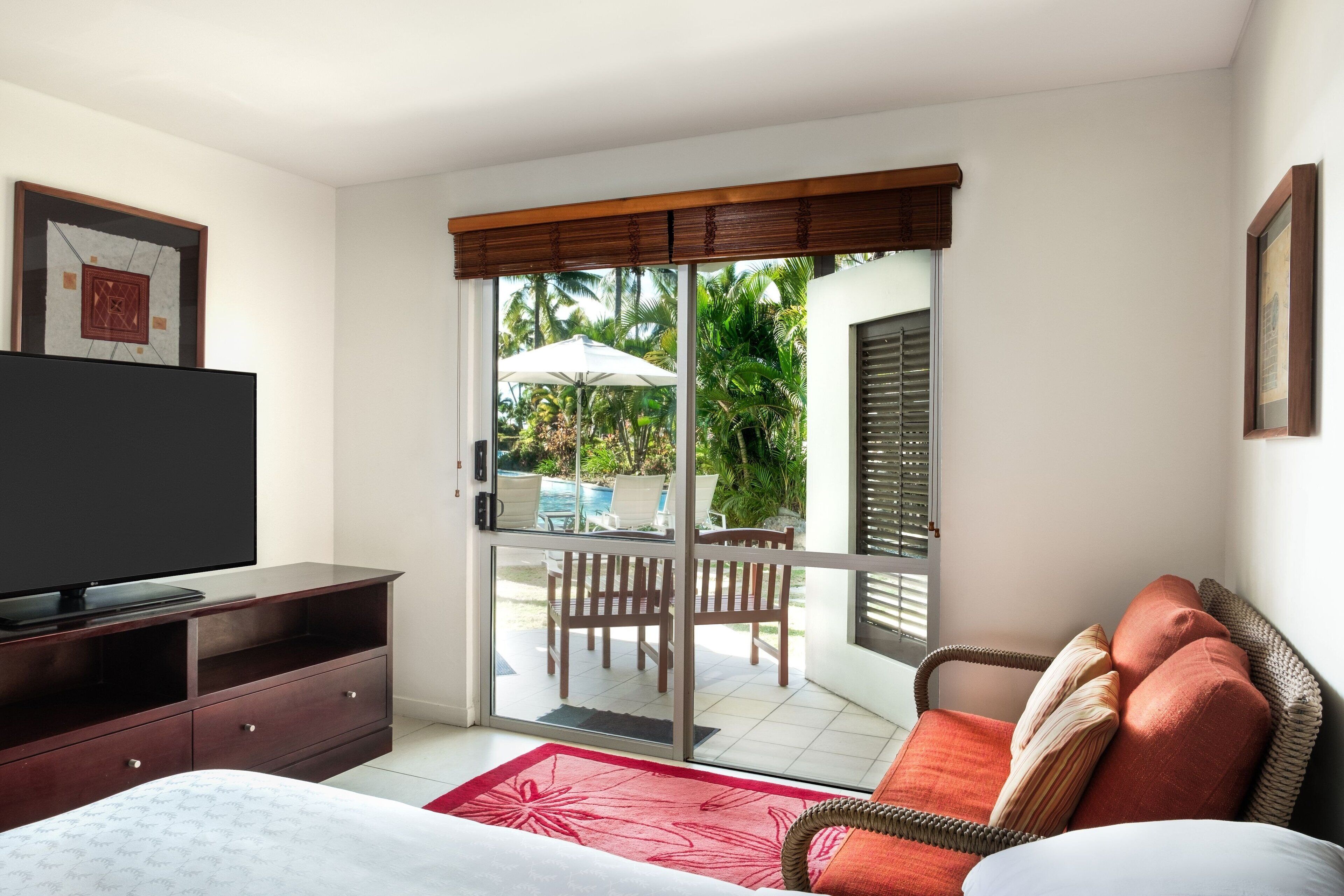 A bedroom with a TV, a bed, a sofa, a sliding glass door to a balcony, and a dining area with chairs and an umbrella.