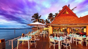 A wooden deck with tables and chairs, overlooking a body of water, with a building featuring a triangular roof and palm trees in the background.