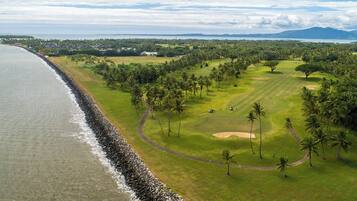 Un campo de golf costero con costa rocosa, palmeras y una cadena montañosa distante.