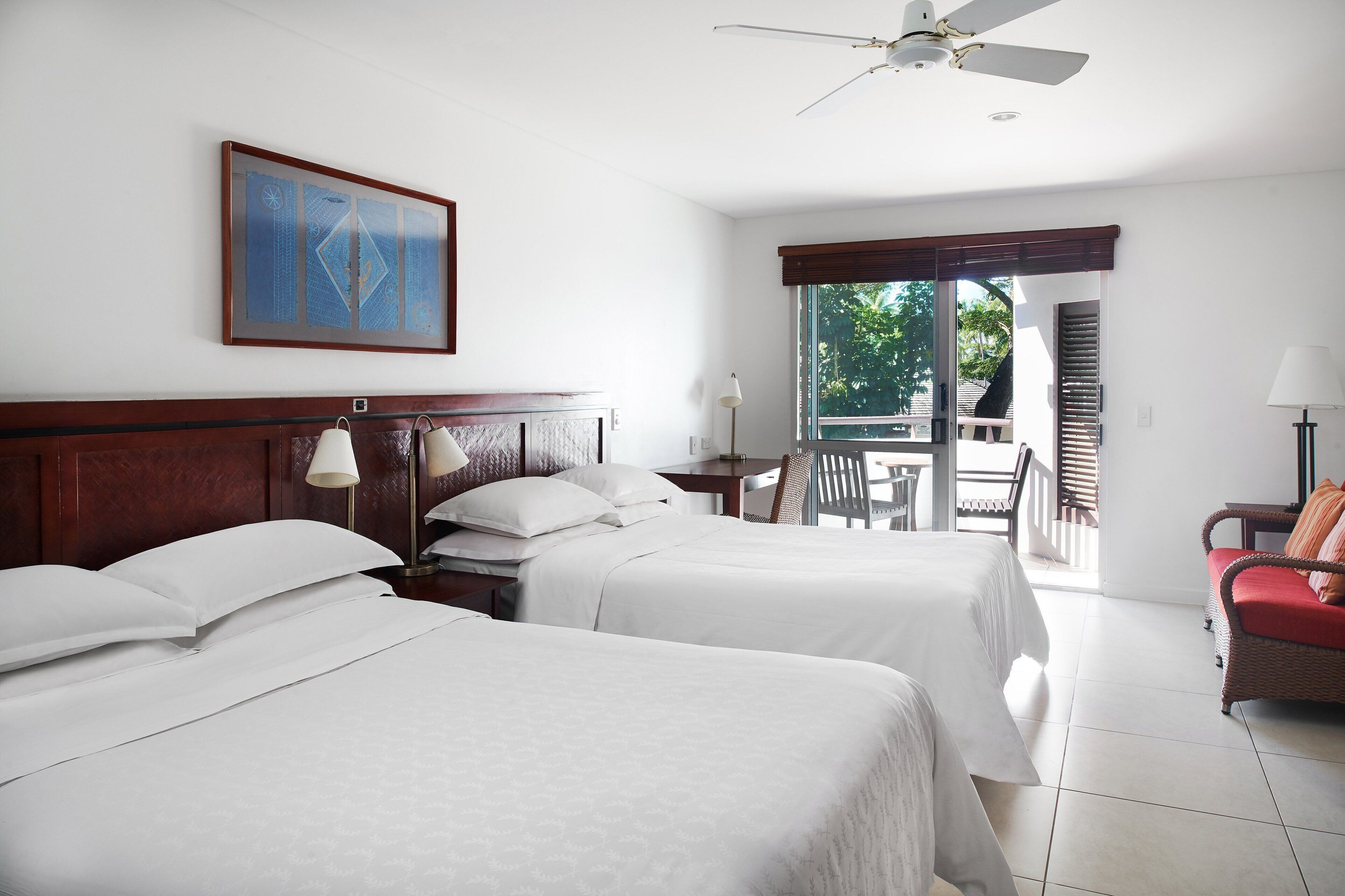 A hotel room with two beds, a ceiling fan, a painting, a desk with a lamp, and a view of the outdoors.