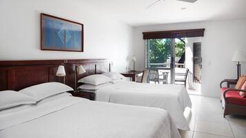 A hotel room with two beds, a ceiling fan, a painting, a desk with a lamp, and a view of the outdoors.