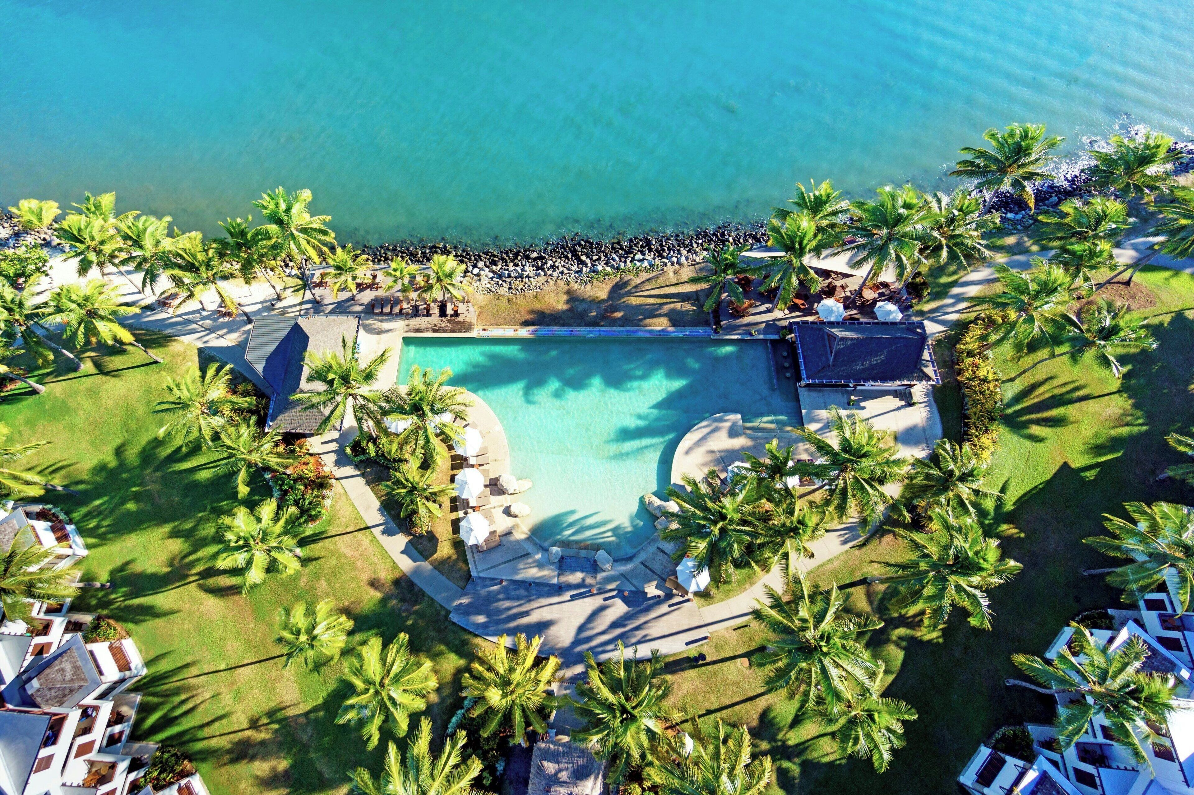 An aerial view of a resort with a central swimming pool, surrounded by palm trees and lounge areas.