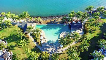 An aerial view of a resort with a central swimming pool, surrounded by palm trees and lounge areas.