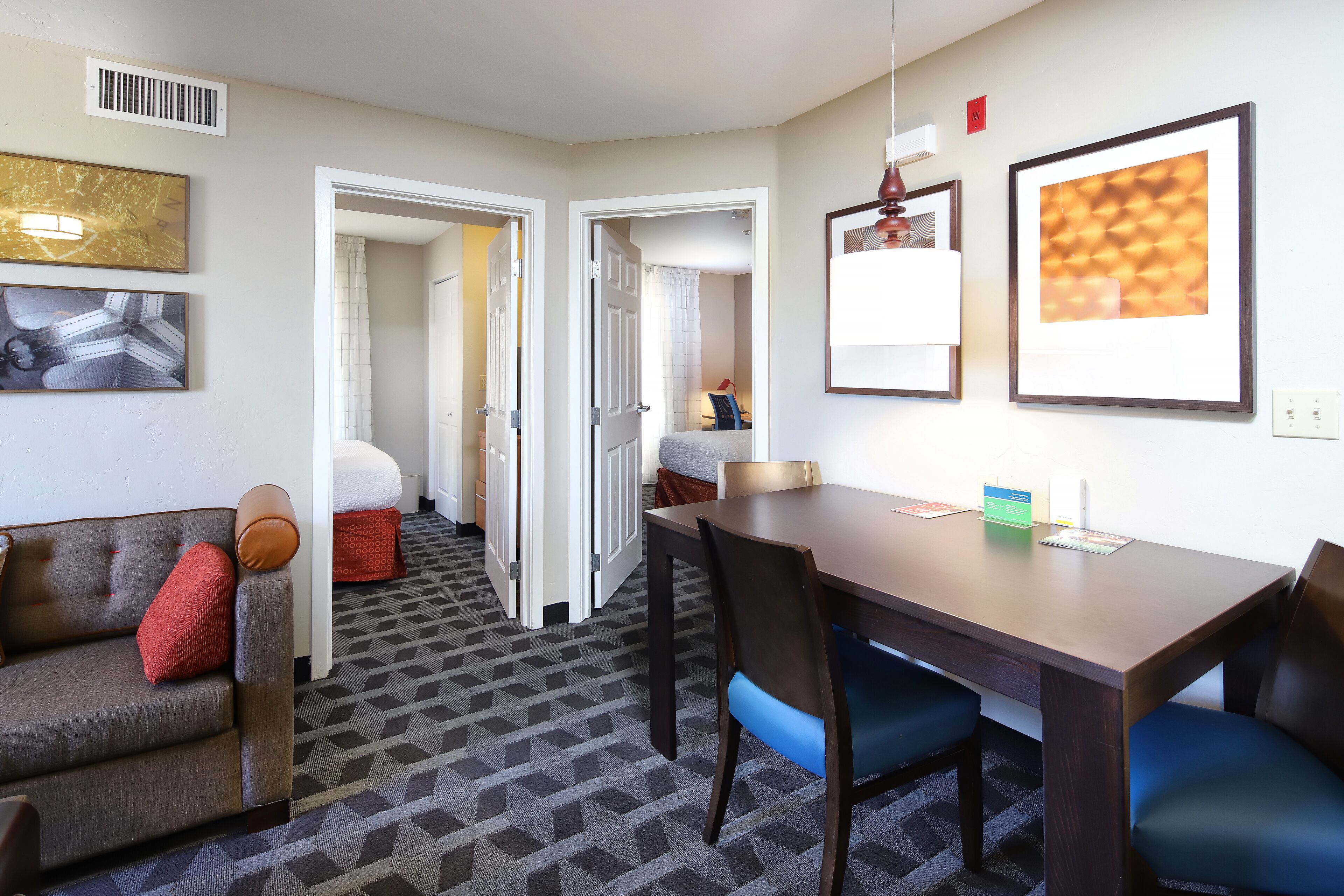 A hotel room with a sofa, a dining table with chairs, a patterned carpet, and framed artwork on the walls.