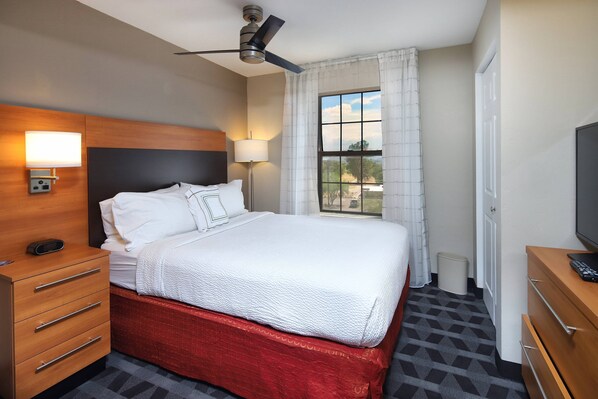 A hotel room with a bed, a wooden nightstand, a lamp, a ceiling fan, a window with curtains, and a TV on a wooden dresser.
