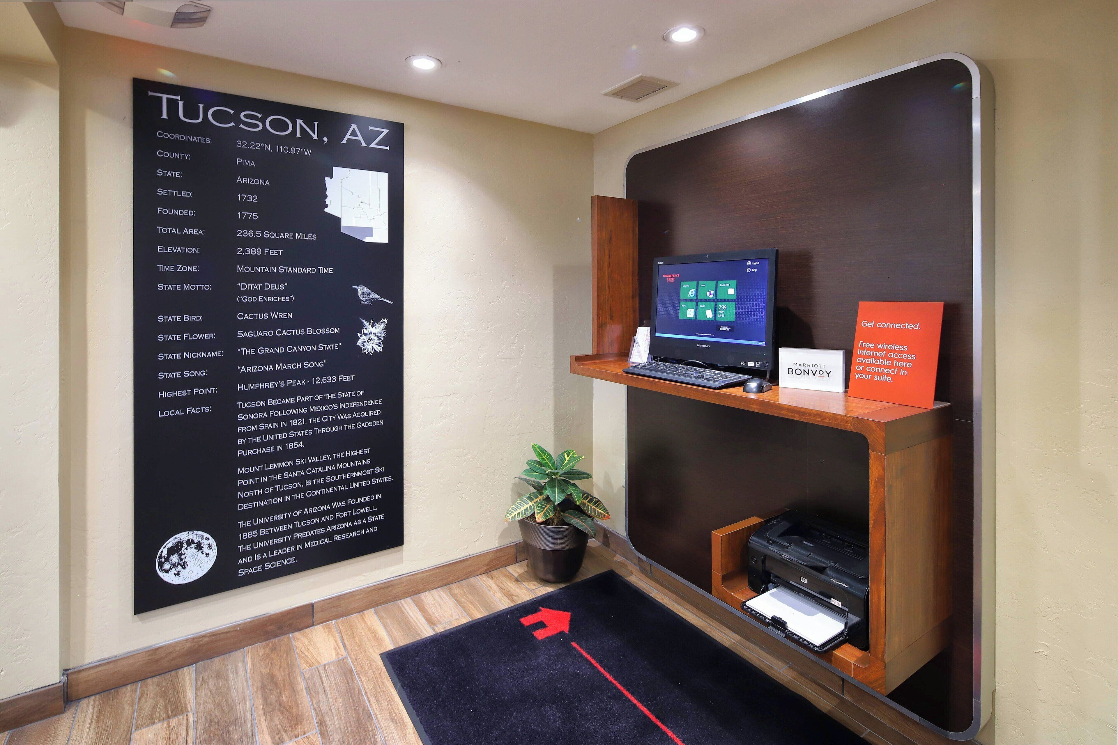 A hotel reception area with a large informational sign about Tucson, AZ, a computer on a shelf, a printer, and a directional arrow on the floor.