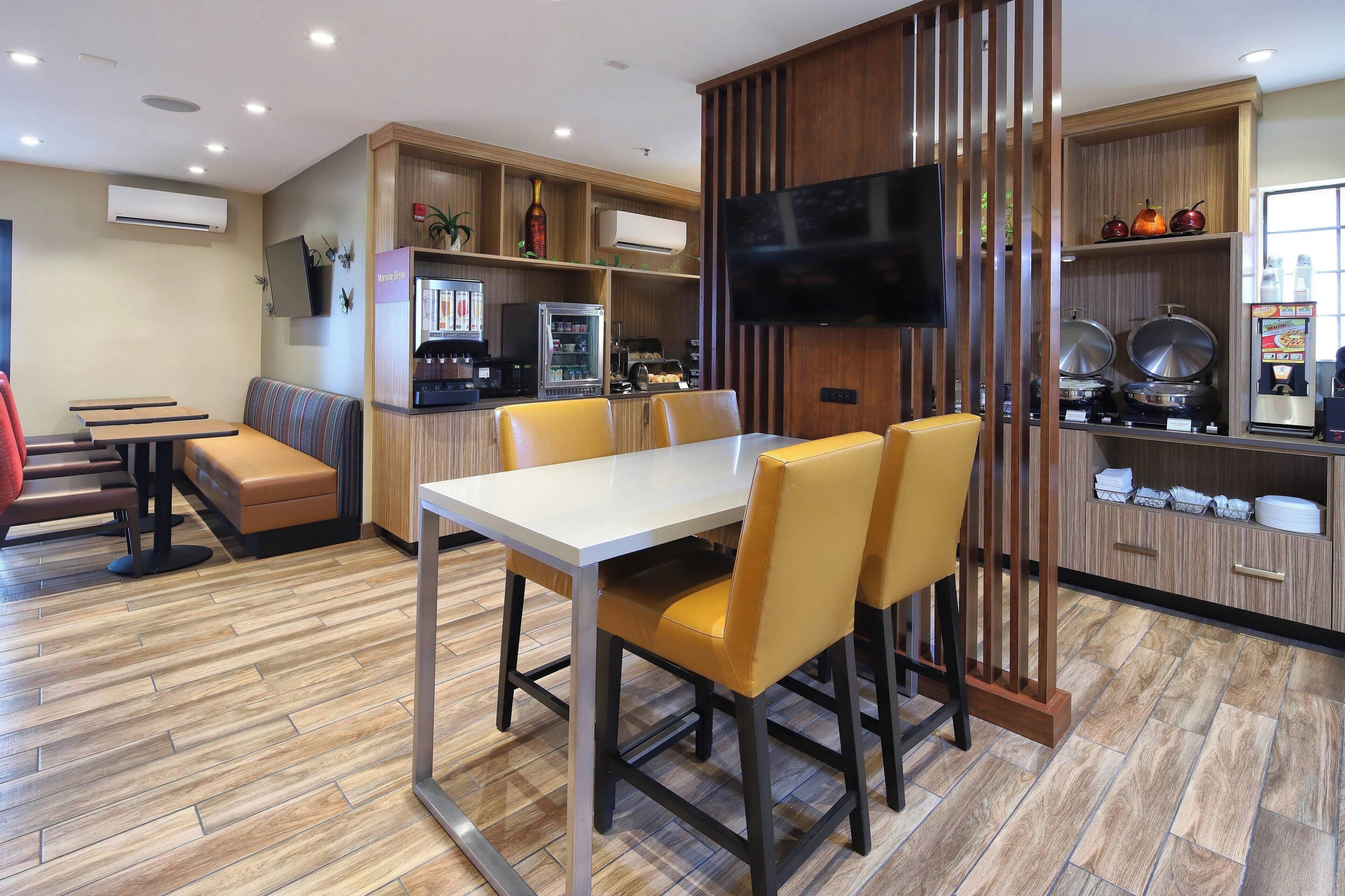 Un comedor moderno con una mesa blanca y sillas amarillas, una zona de estar con un banco, y una cocina con un televisor y diversos electrodomésticos.