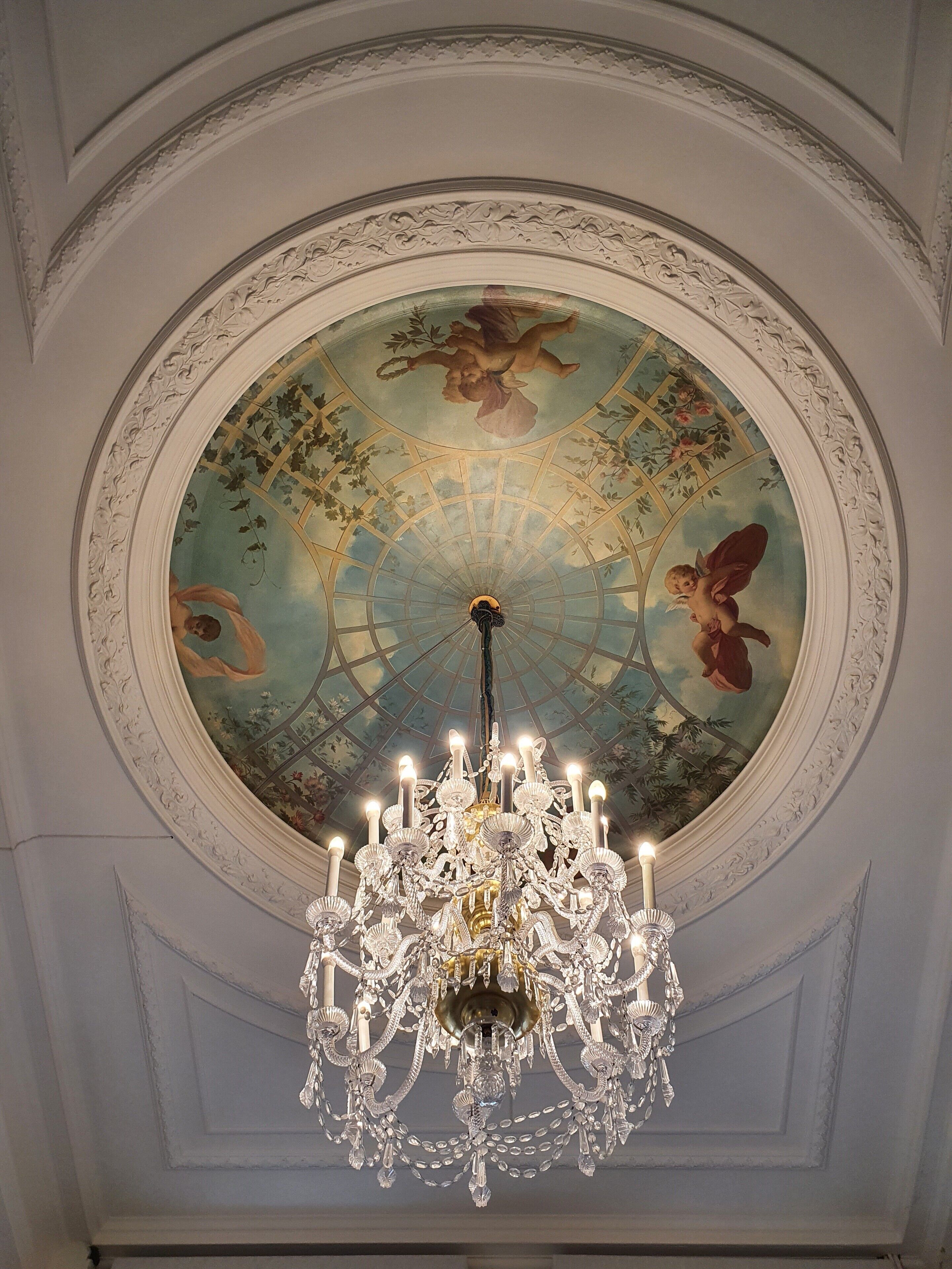 A ceiling with a detailed fresco, a large ornate chandelier, and a circular architectural design.
