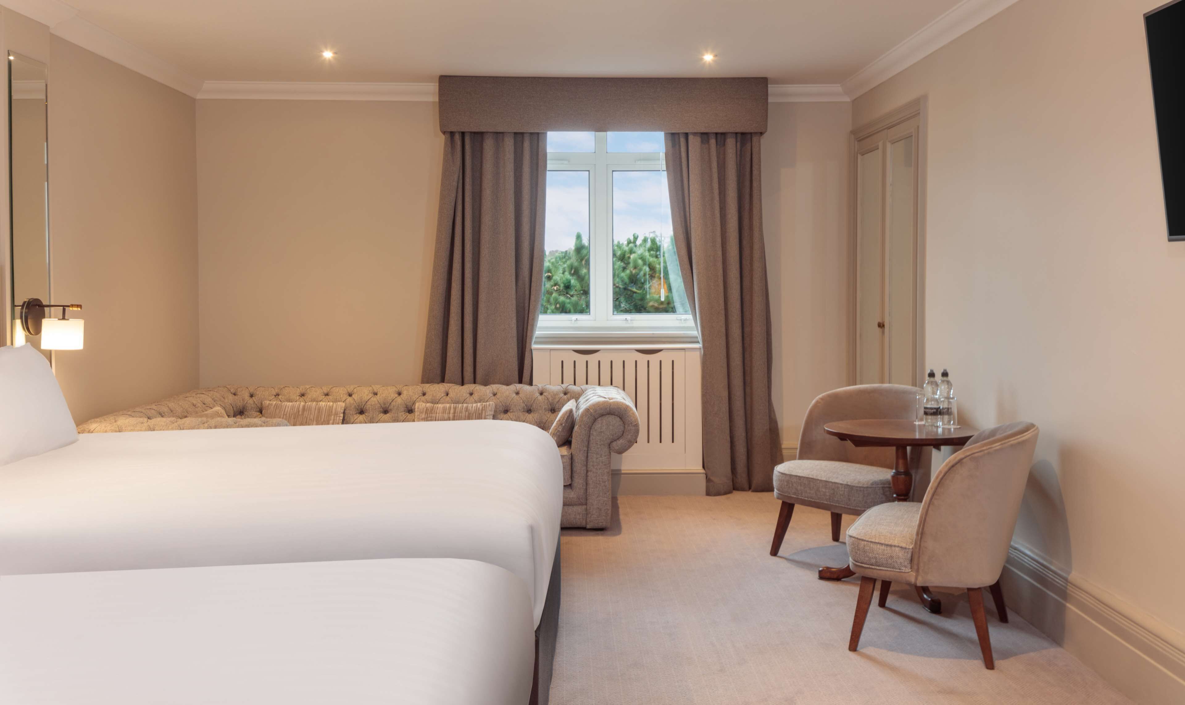 A hotel room with two beds, a sitting area with a table and chairs, a window with curtains, and a radiator.