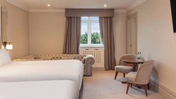 A hotel room with two beds, a sitting area with a table and chairs, a window with curtains, and a radiator.