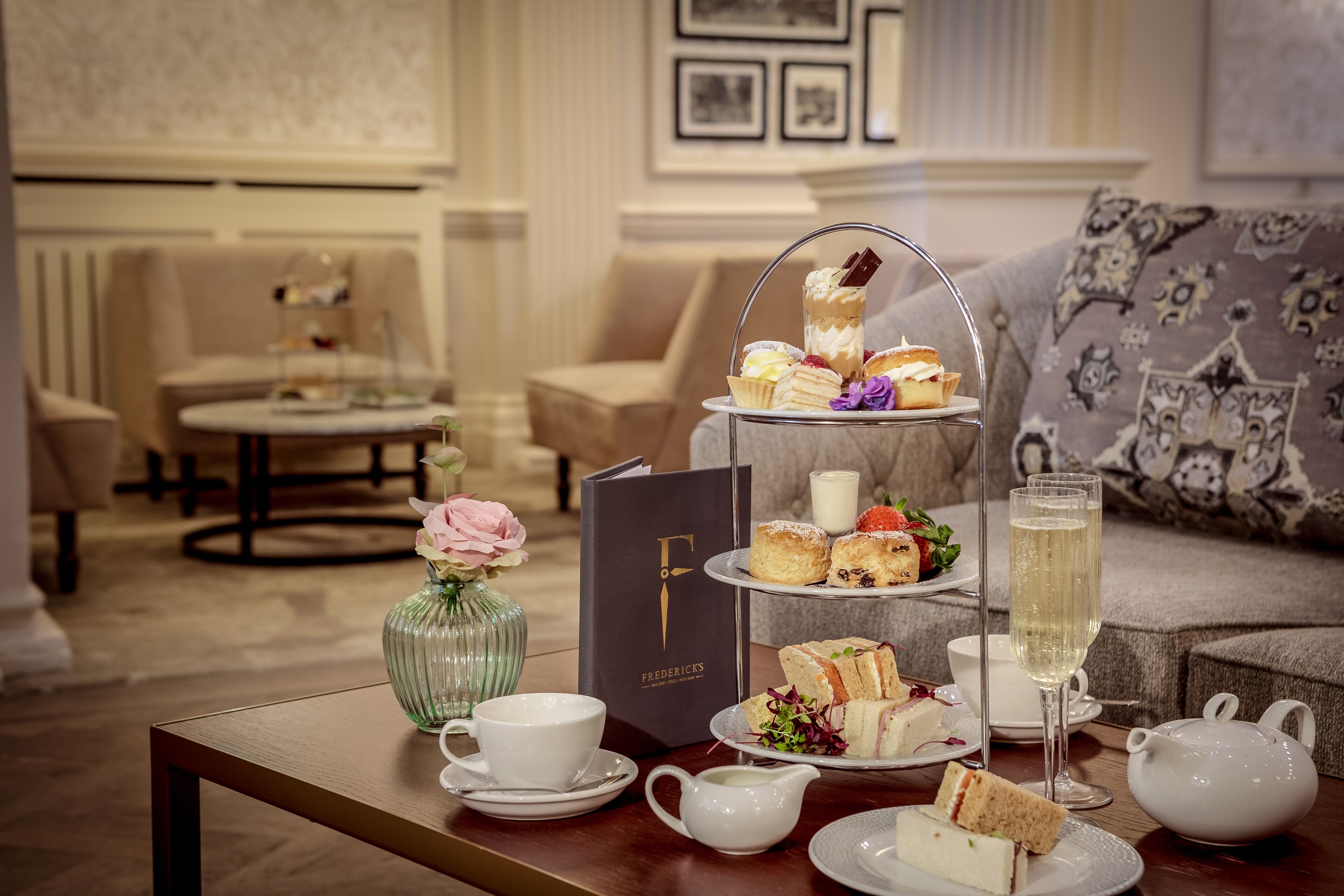 A tea service setup with sandwiches, scones, and a teapot on a table in a well-appointed room with a sofa and framed pictures on the wall.