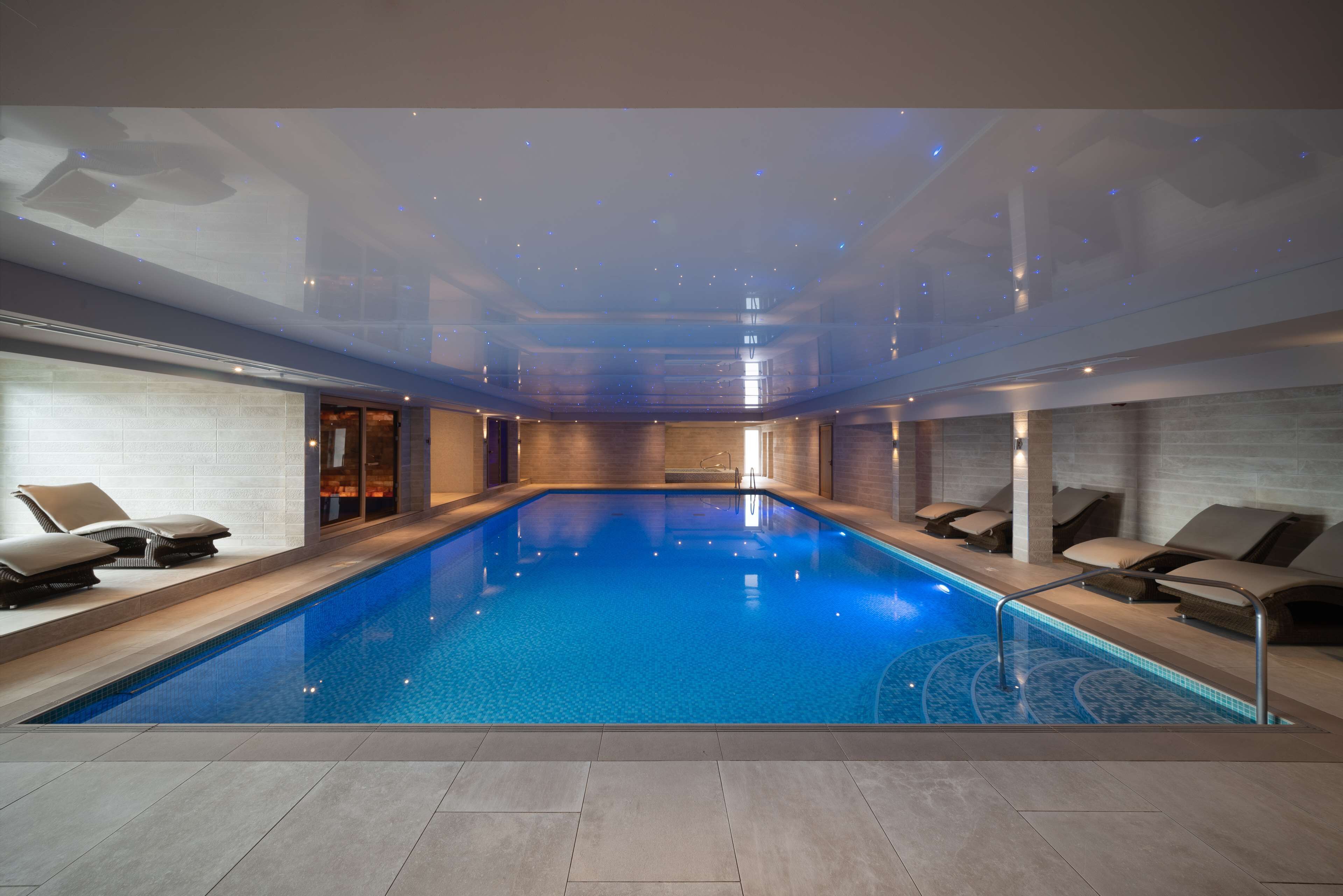 A modern indoor swimming pool with clear blue water, surrounded by lounge chairs and a glass ceiling.