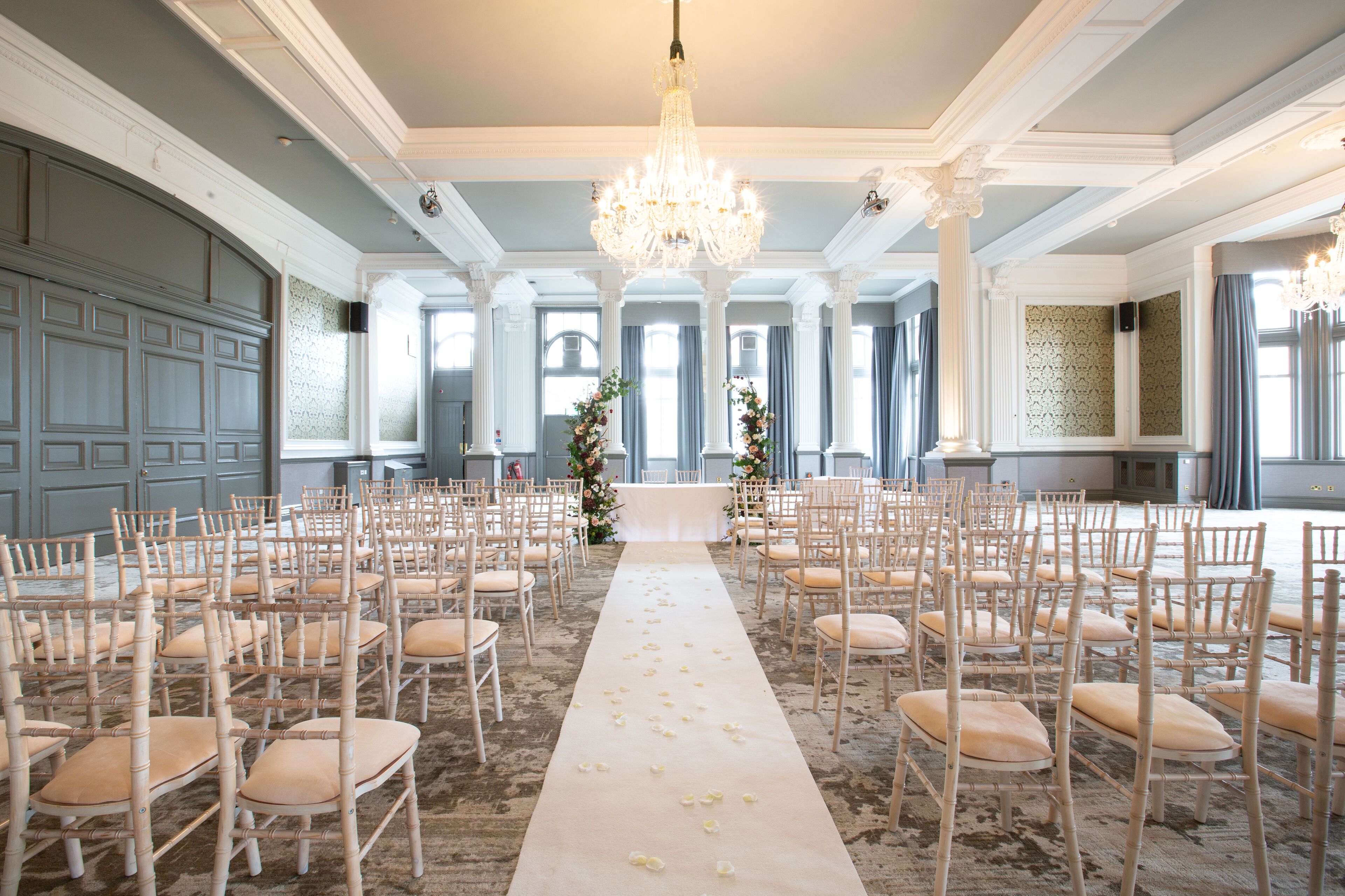 A grand hall with rows of wooden chairs, a central aisle with flower petals, and a chandelier.