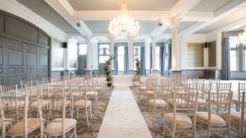 A grand hall with rows of wooden chairs, a central aisle with flower petals, and a chandelier.