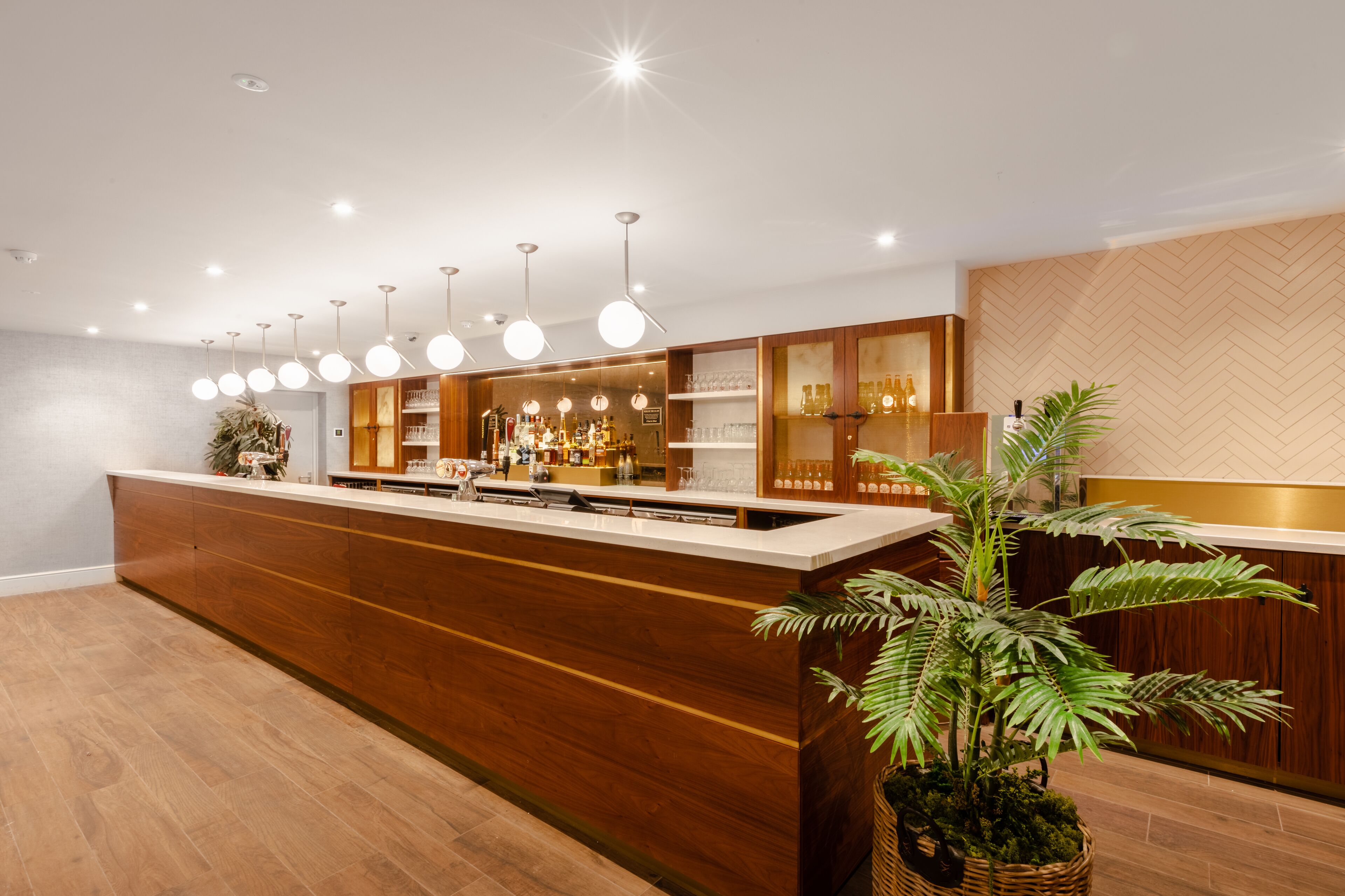 A modern bar area with a wooden counter, hanging lights, and a potted plant.
