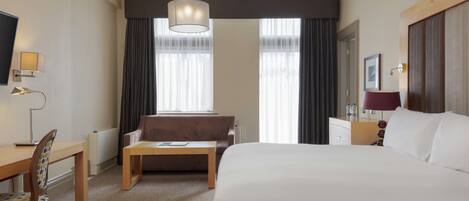 A hotel room with a large bed, a desk with a chair, a sofa, a coffee table, and a TV.