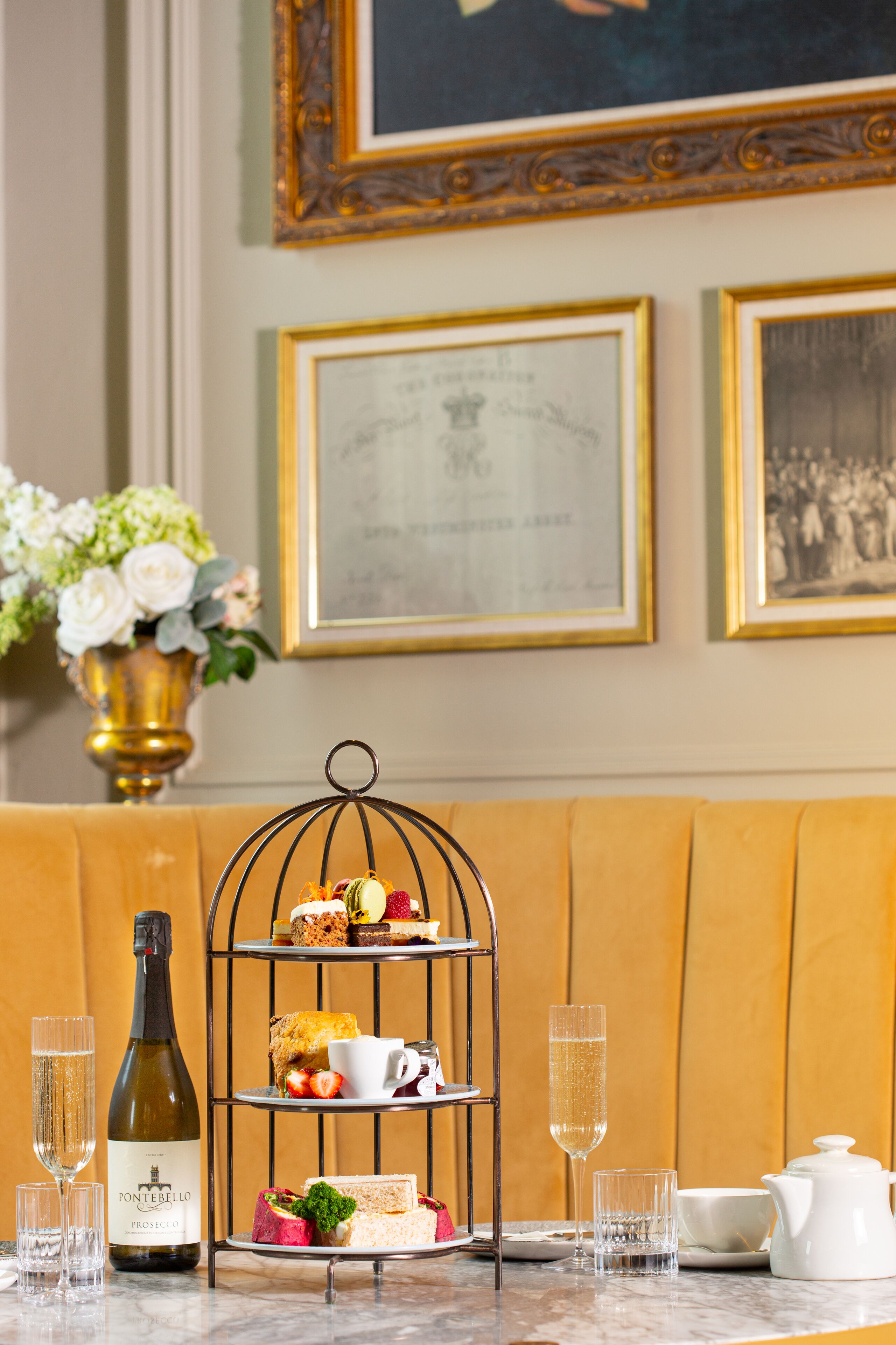 A dining table with a bottle of wine, champagne glasses, a tiered dessert stand, and a white teapot.