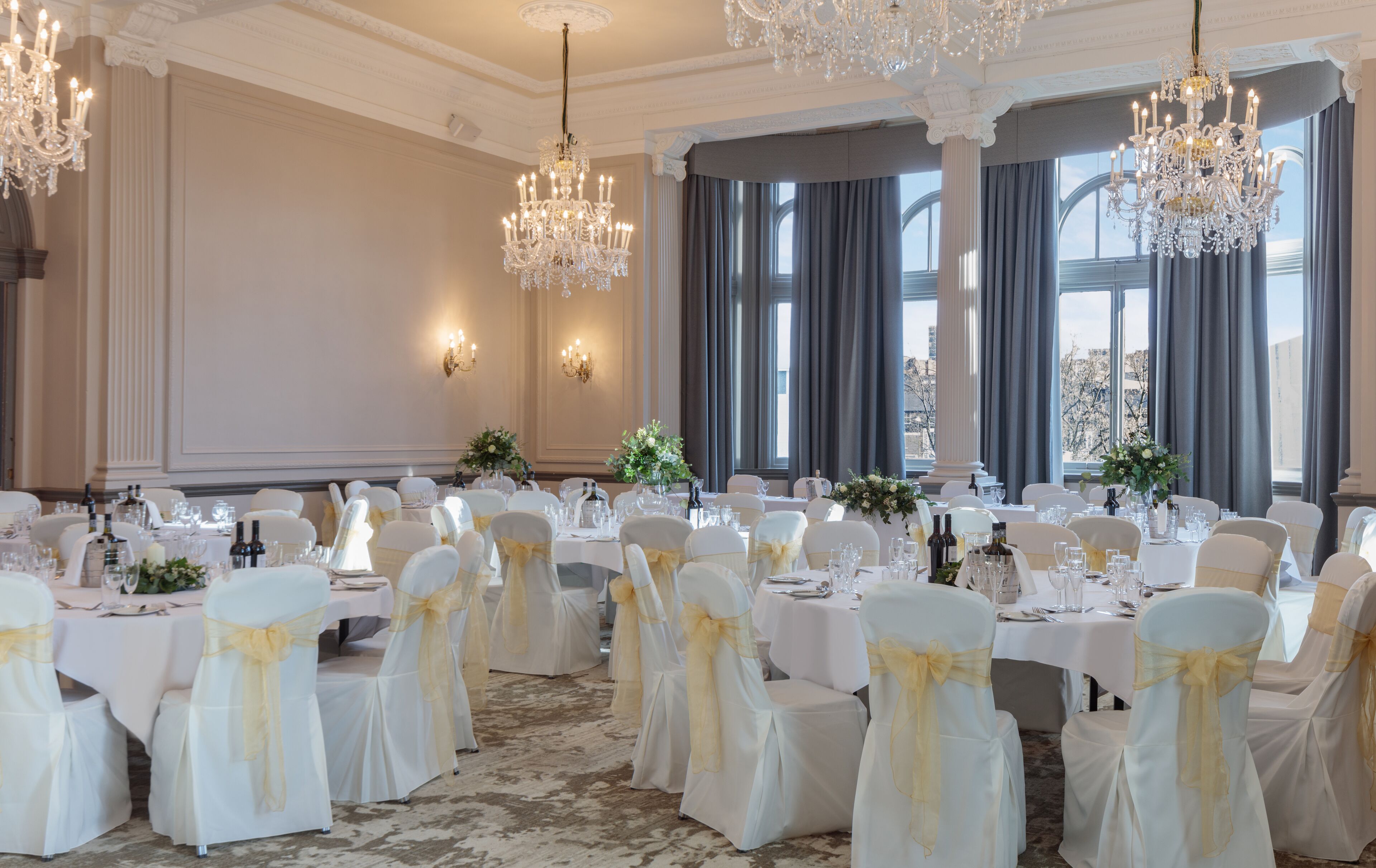 A banquet hall with round tables, white chairs with yellow sashes, chandeliers, and large windows with curtains.